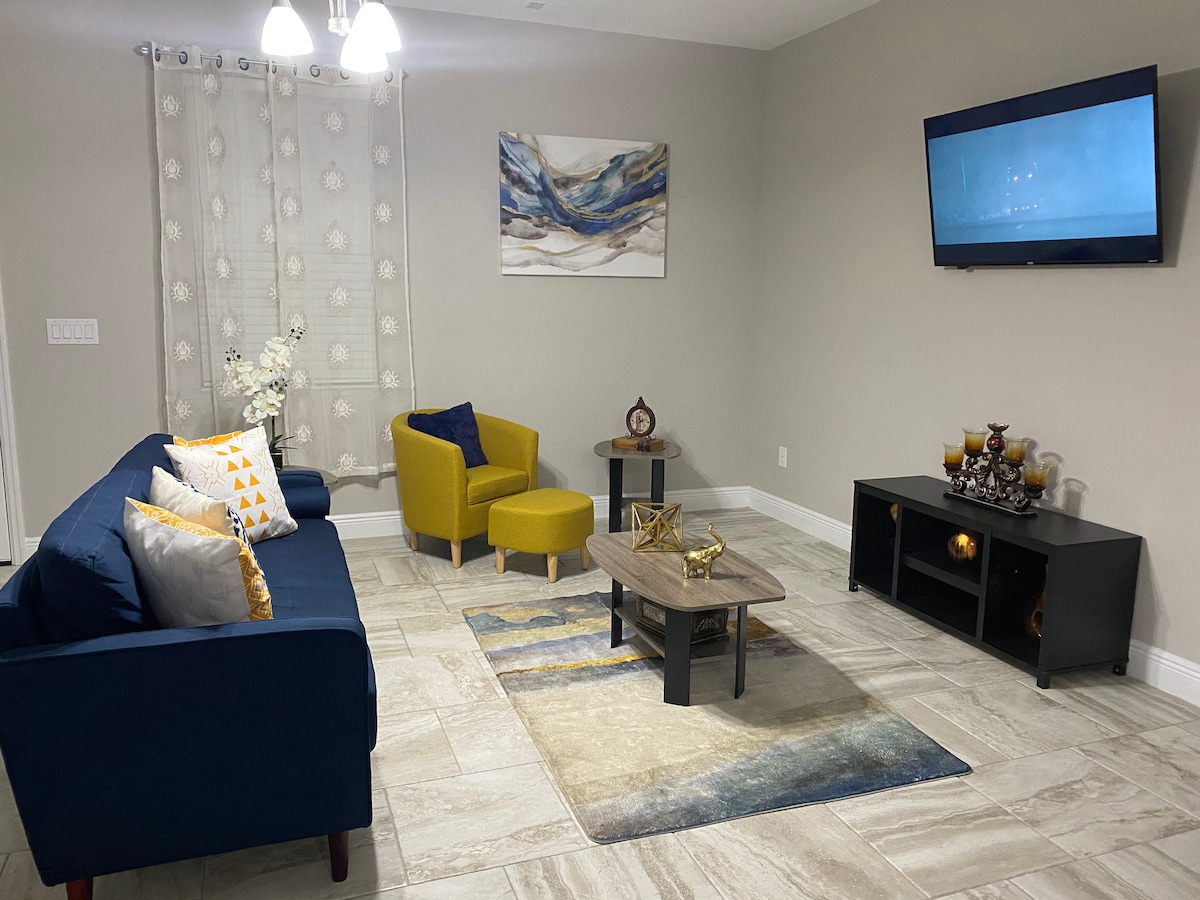 A modern living area is furnished with a blue sofa adorned with decorative pillows and a yellow accent chair. A coffee table sits on a patterned rug, and a media console holds decorative items. A wall-mounted television is visible, with soft lighting provided by a ceiling fixture.