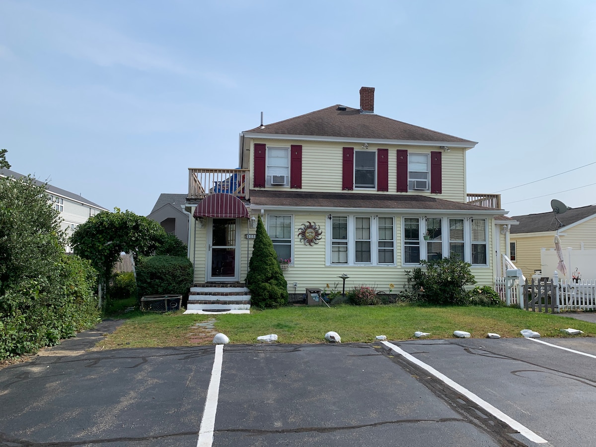 A two-story yellow house is displayed with red shutters and a welcoming front entrance. Lush greenery surrounds the property, and a small staircase leads to a covered porch. A designated parking area is visible in the foreground, accommodating off-street vehicle access.