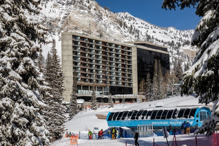 A modern building is visible amidst a snowy landscape, surrounded by tall trees. The structure features multiple levels with large windows. A ski lift is positioned nearby, with skiers preparing to embark on their journey. Snow-covered mountains rise majestically in the background.