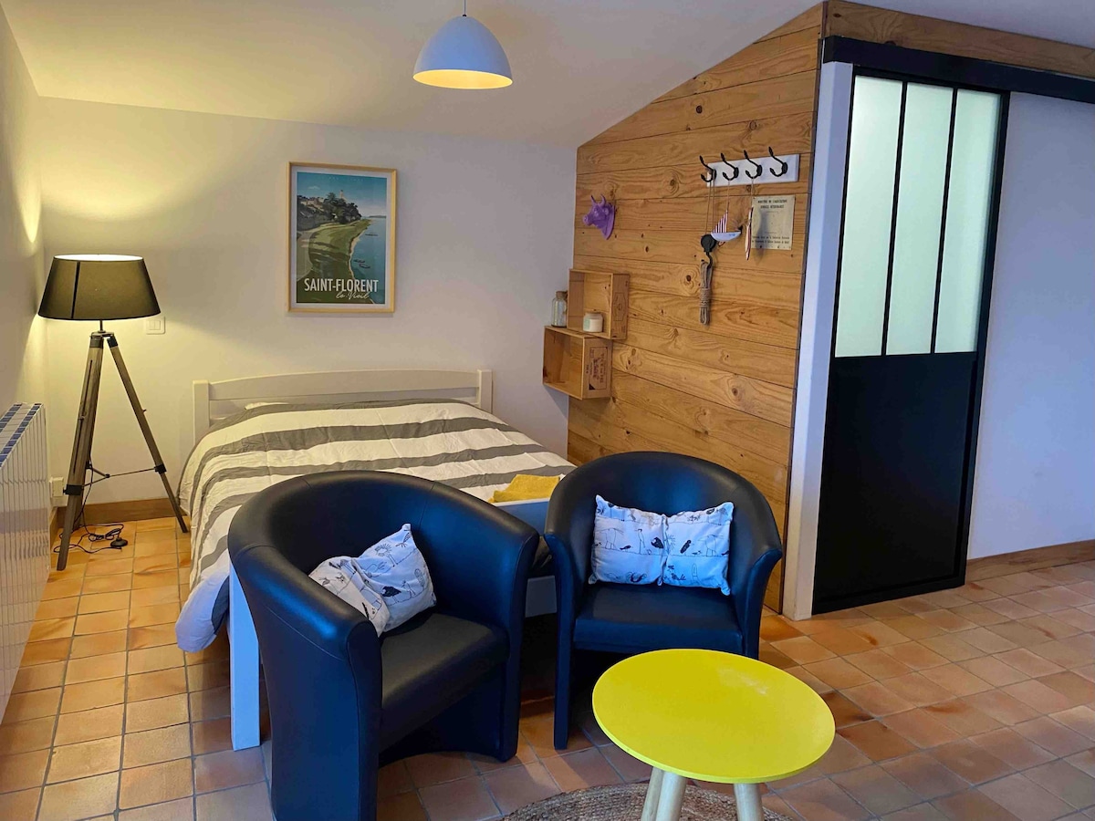 A living area is presented with a spacious bed against a wooden accent wall, complemented by a framed artwork. Two black armchairs with decorative cushions are positioned near a yellow table. Soft lighting is provided by a floor lamp, creating a cozy ambiance.