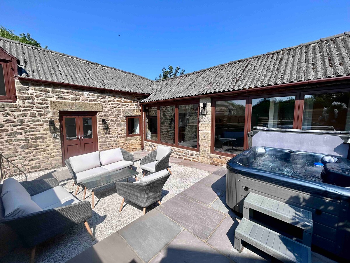 An enclosed private patio area features a hot tub, surrounded by comfortable seating arrangements. The property showcases a blend of natural stone and glass, with large windows allowing sunlight to fill the space. The tranquil outdoor setting invites relaxation amid nature.