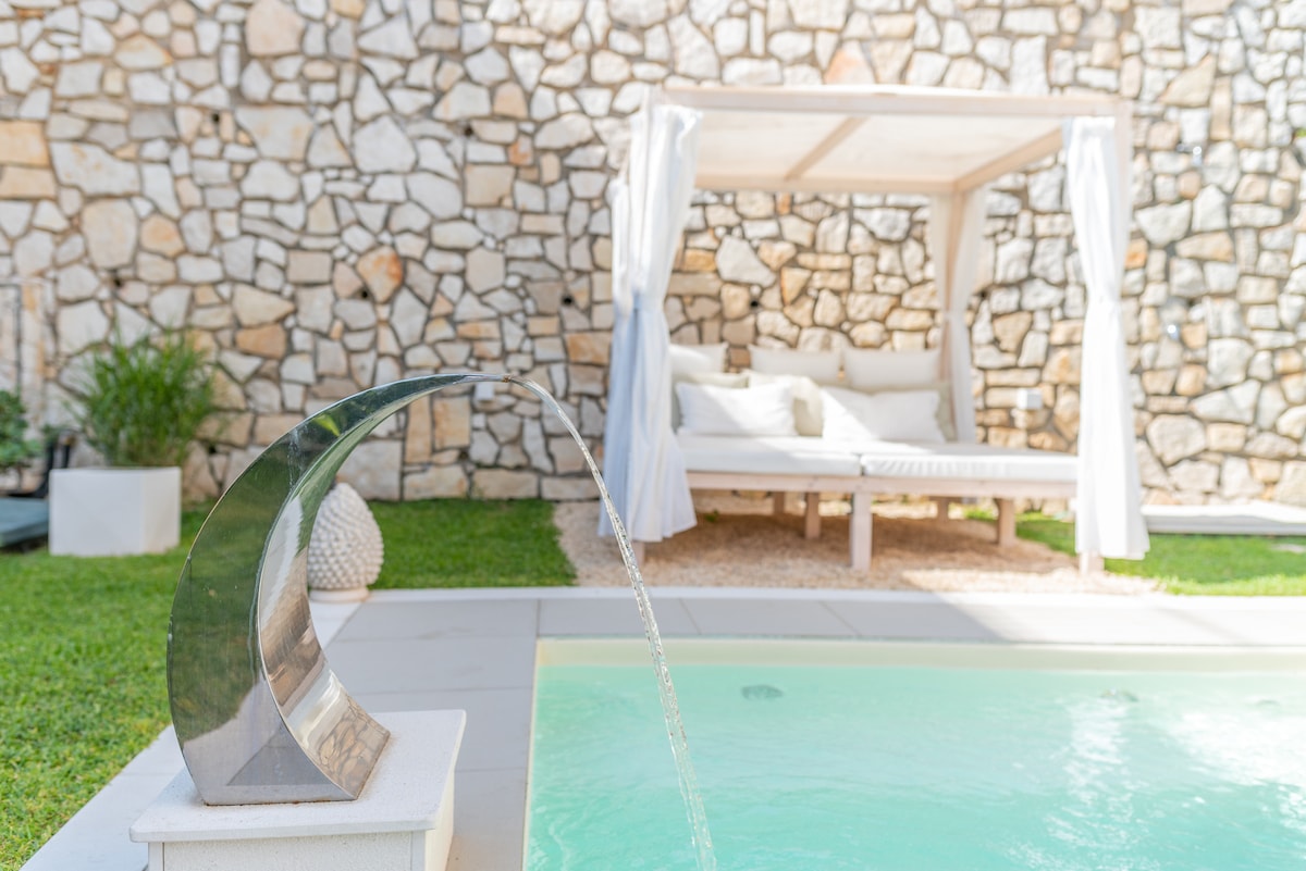 A private mini pool is showcased with a gentle water feature flowing from a sculptural spout. A shaded daybed sits nearby, draped with white curtains, surrounded by lush greenery, creating a serene outdoor space for relaxation.