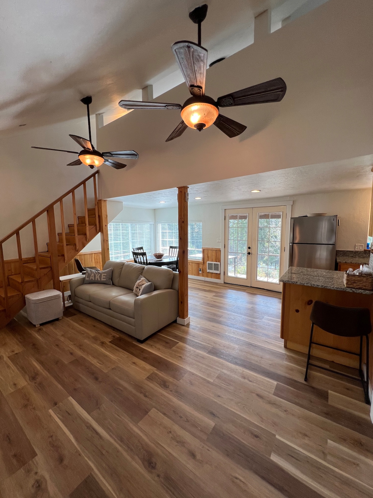The interior space features an open floor plan, showcasing a light-filled living room with a comfortable sofa. Two ceiling fans provide airflow. A staircase leads to the upper loft, while a dining area adjacent to modern kitchen appliances is visible through the large windows.