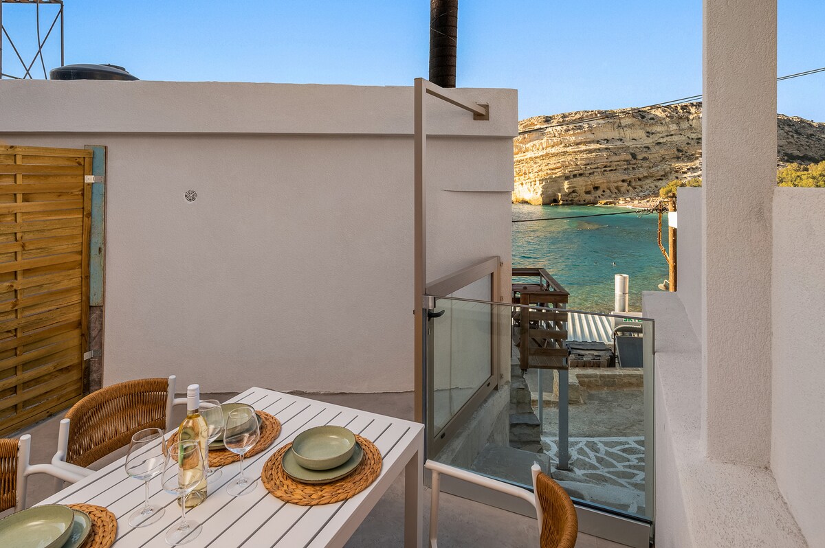 A private terrace is featured, showcasing a dining table set for four, including plates and glassware. The view reveals water and rocky cliffs in the background, framed by the blue sky. Natural light brightens the space, enhancing the serene outdoor environment.