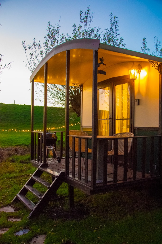 A charming vintage caravan features a welcoming porch with wooden steps leading up. Two wicker chairs sit on the porch, illuminated by a soft lamp, with a barbecue grill nearby. A serene, grassy landscape is visible in the background, creating a peaceful ambiance.