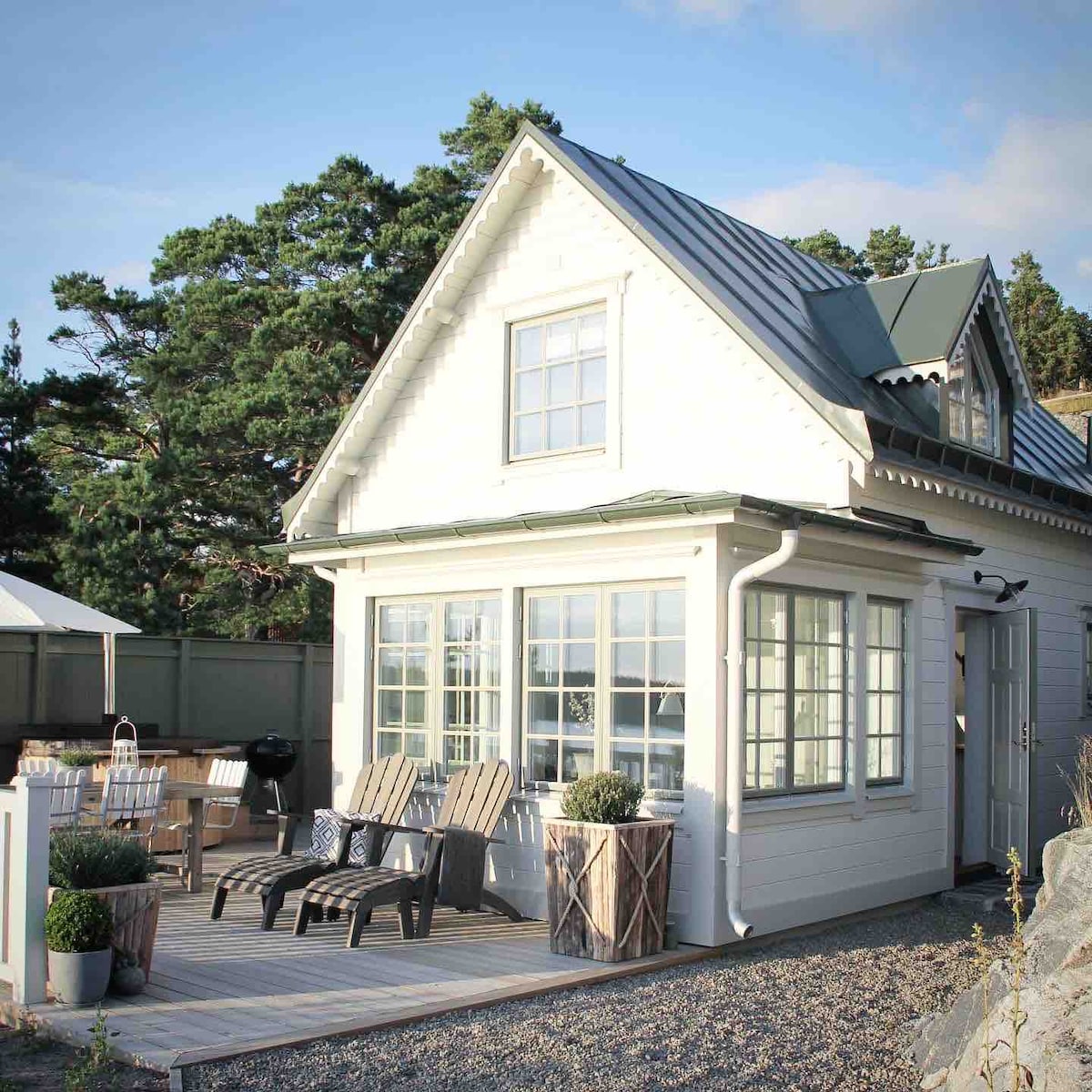 A charming white house features large windows, allowing natural light to fill the interior. Outdoor seating arrangements are visible on a wooden deck, surrounded by neatly trimmed plants in decorative containers. The sloped roof and wooden details convey a traditional style.