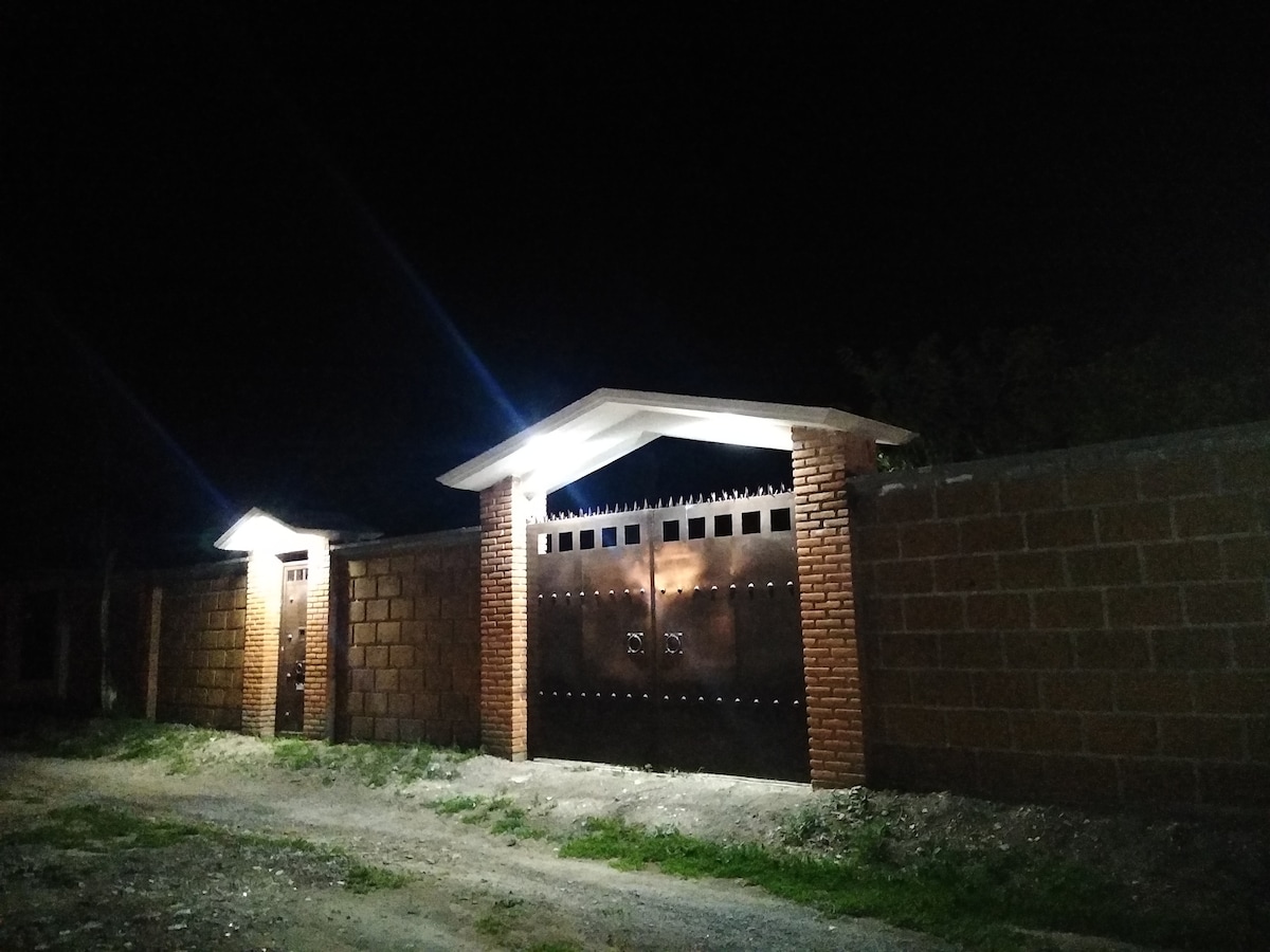 The entrance features a sturdy brick wall illuminated by soft lights, guiding guests to the large, metallic gates. The inviting pathway is lined with grass, creating a welcoming approach to the property.
