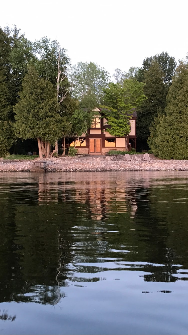 A contemporary lake house is depicted from the water, surrounded by tall trees and greenery. The structure features large windows reflecting the calm water, with a prominent entrance flanked by natural stone elements along the shoreline.