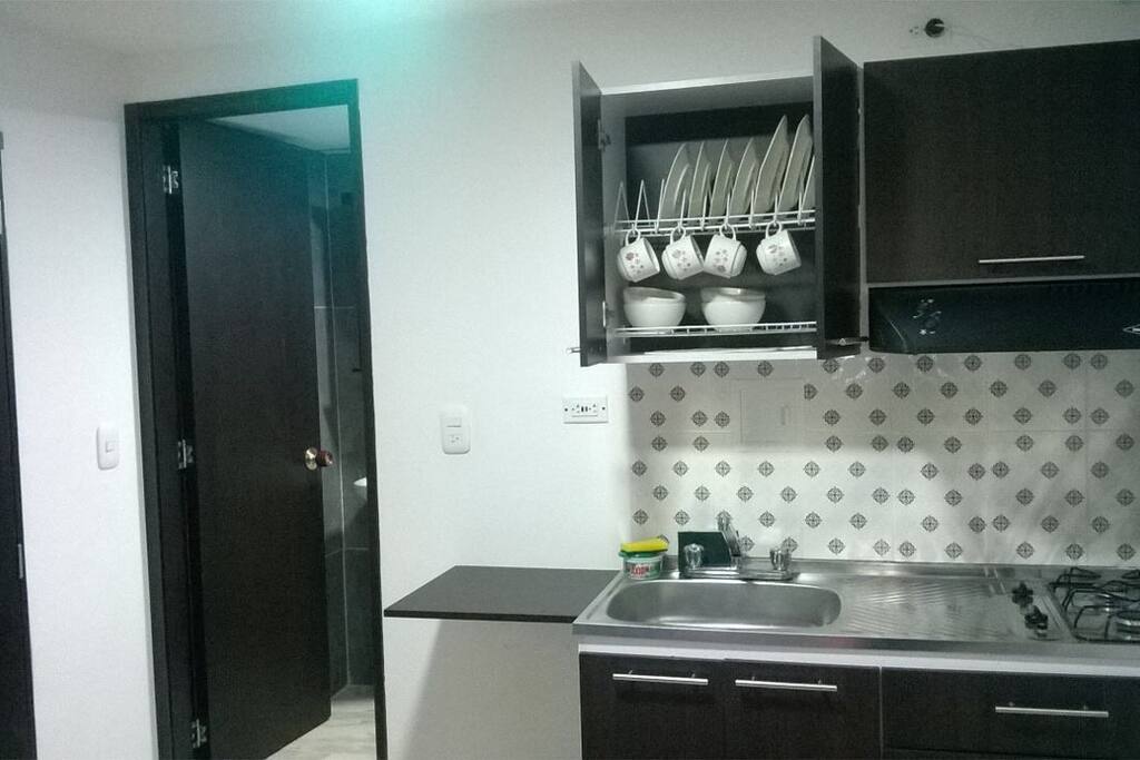 Cocina con juego completo de platos, ollas, cubiertos y nevera. La puerta es la entrada del baño