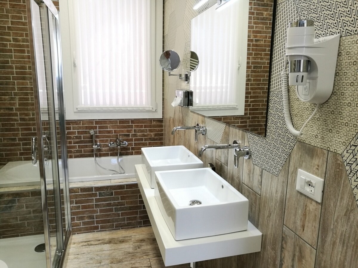 The bathroom is designed with modern fixtures and features a dual sink vanity with sleek white basins. A spacious shower stall and a bathtub are visible, surrounded by a stylish mix of brick and patterned tiles. Natural light filters through the window with blind coverings.