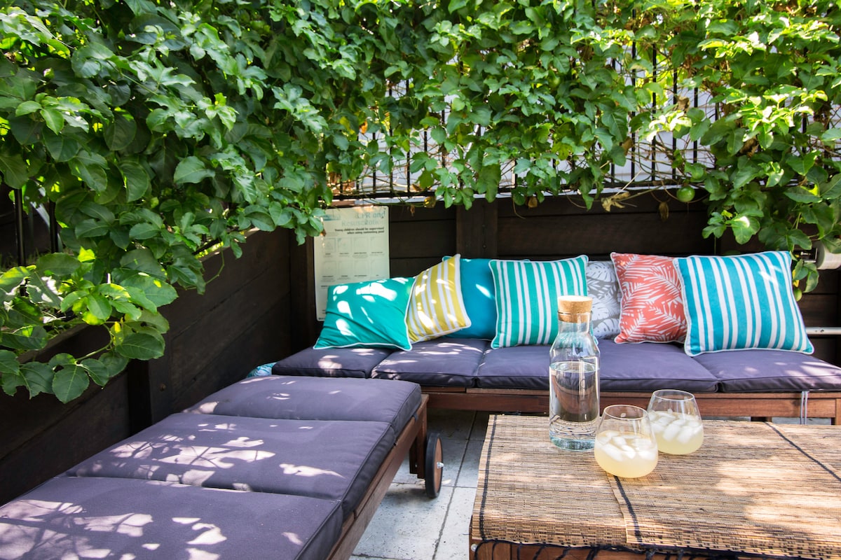 A relaxing outdoor seating area is surrounded by lush greenery. Comfortable lounge chairs are adorned with colorful cushions. A wooden table holds a pitcher of refreshing beverage and two glasses, enhancing the tranquil atmosphere for enjoying the outdoors.