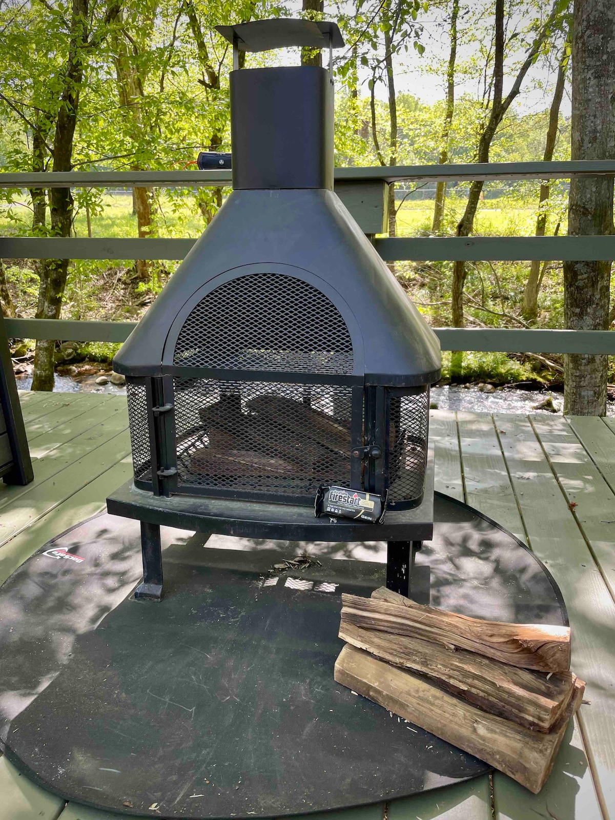 A modern outdoor fireplace is set on a circular platform, accompanied by a stack of firewood. The surrounding area features green trees and a serene creek, creating a peaceful environment for relaxation.