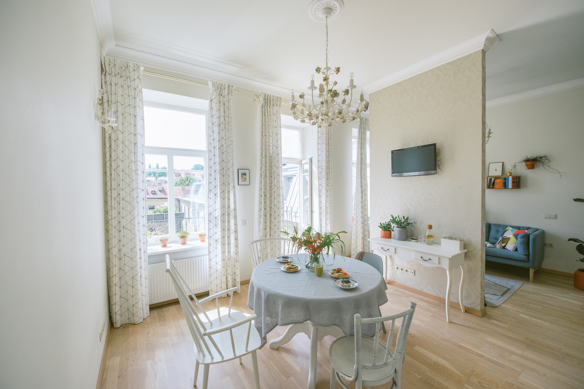 A dining area is highlighted by a round table set with dishes, surrounded by white chairs. Natural light enters through large windows adorned with light curtains, casting a warm glow. A chandelier hangs above, complementing a TV mounted on the wall, while plants add a touch of greenery.