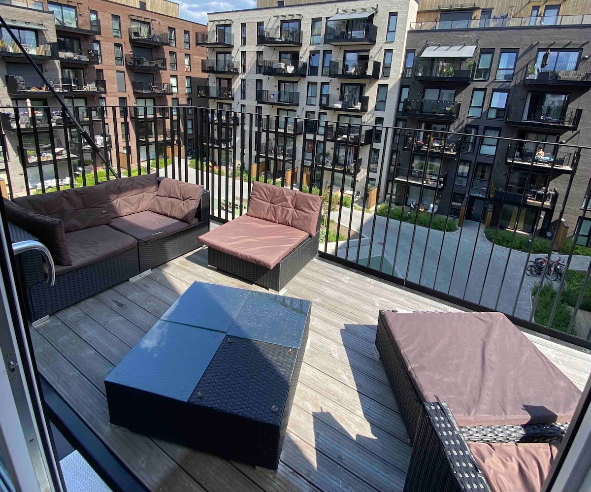An outdoor balcony features comfortable seating arrangements with brown cushions, accompanied by a glass-topped table. The view overlooks a landscaped courtyard with multiple floors of residential buildings and various balconies in the background.