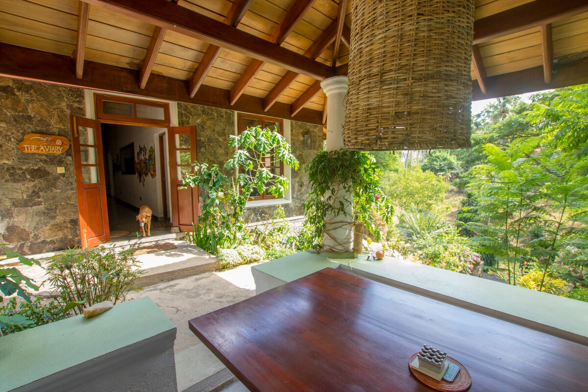 A covered porch is adorned with natural stone walls and a wooden ceiling, featuring a large table. Lush greenery and ornamental plants surround the area, creating a serene setting. A glimpse of an inviting entryway can be seen in the background.