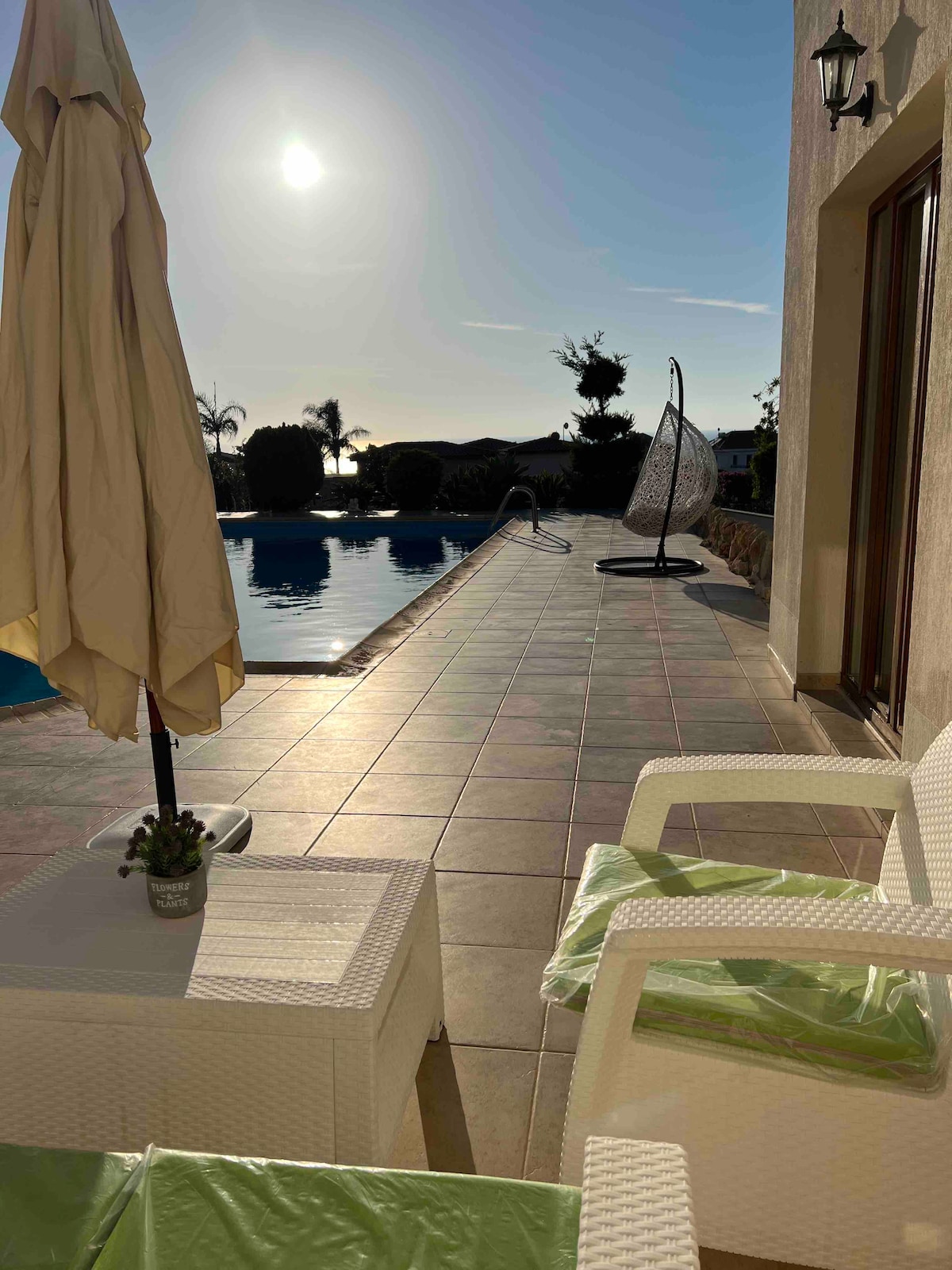 A sunlit patio area is presented, featuring a swimming pool that reflects the warm glow of the sun. White lounge chairs are arranged beside the pool, accompanied by a small table and an umbrella. Lush greenery can be seen in the background, enhancing the serene atmosphere.