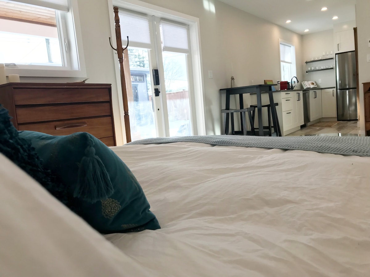 A close-up view of a spacious bed featuring soft white linens and a decorative teal pillow. The background displays a kitchenette with modern appliances and a dining area illuminated by natural light from nearby glass doors.
