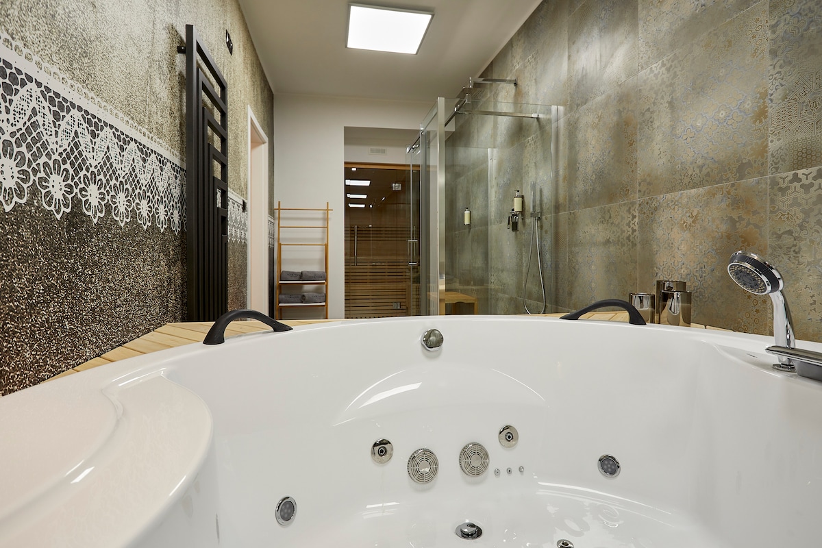 A modern spa area features a white jacuzzi with multiple jets. The backdrop includes tiled walls with intricate designs and a glass shower area. Wooden accents and a sauna can be seen in the distance, contributing to a relaxing environment.