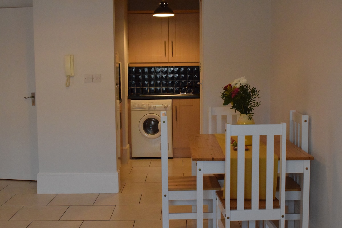 A bright dining area features a wooden table surrounded by white chairs. A compact kitchenette with light wood cabinetry is visible in the background, equipped with a washing machine. A vase of flowers adds a pop of color to the table, creating a welcoming environment.