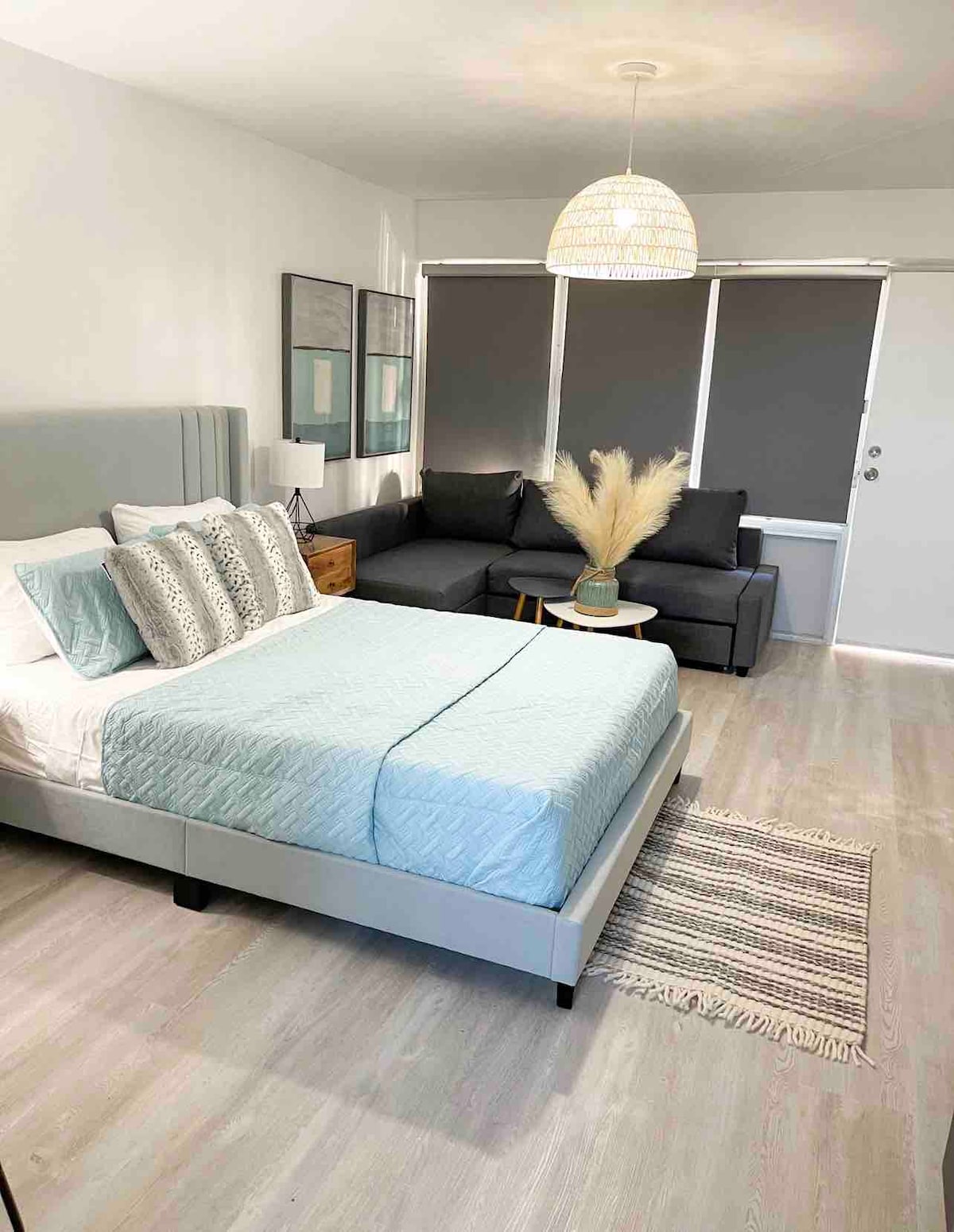 A cozy sleeping area features a spacious bed with a light blue coverlet and decorative pillows. A sectional sofa is positioned nearby, accompanied by a small side table holding a decorative vase with dried grass. Large windows are covered with shades, allowing for flexible light control.