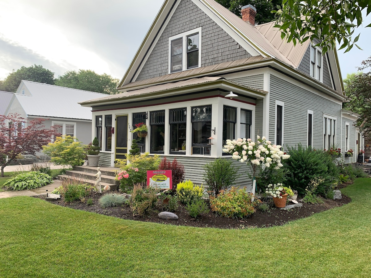 The exterior of a renovated 1910 Craftsman home is showcased, featuring a spacious front porch with multiple windows. Lush greenery and vibrant flowers adorn the well-maintained garden. A welcoming sign is visible in the yard, which is situated on a corner lot with a manicured lawn.