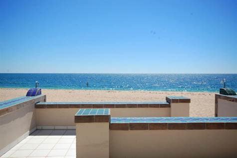 A clear view of the beach and ocean is presented, with sandy shore visible beyond a tiled patio. The horizon offers a seamless blend of blue waters under a cloudless sky, enhancing the tranquil coastal atmosphere.