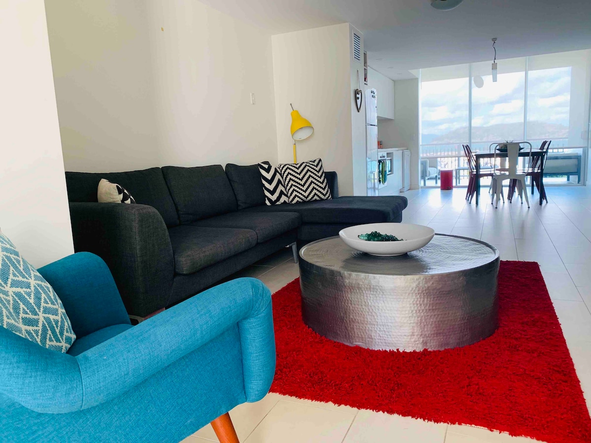A modern living area is highlighted by a sectional sofa and a contrasting blue chair. A round coffee table centers the space, complemented by a vibrant red rug. Large windows provide natural light and an open view towards the dining area, featuring a table set for four.