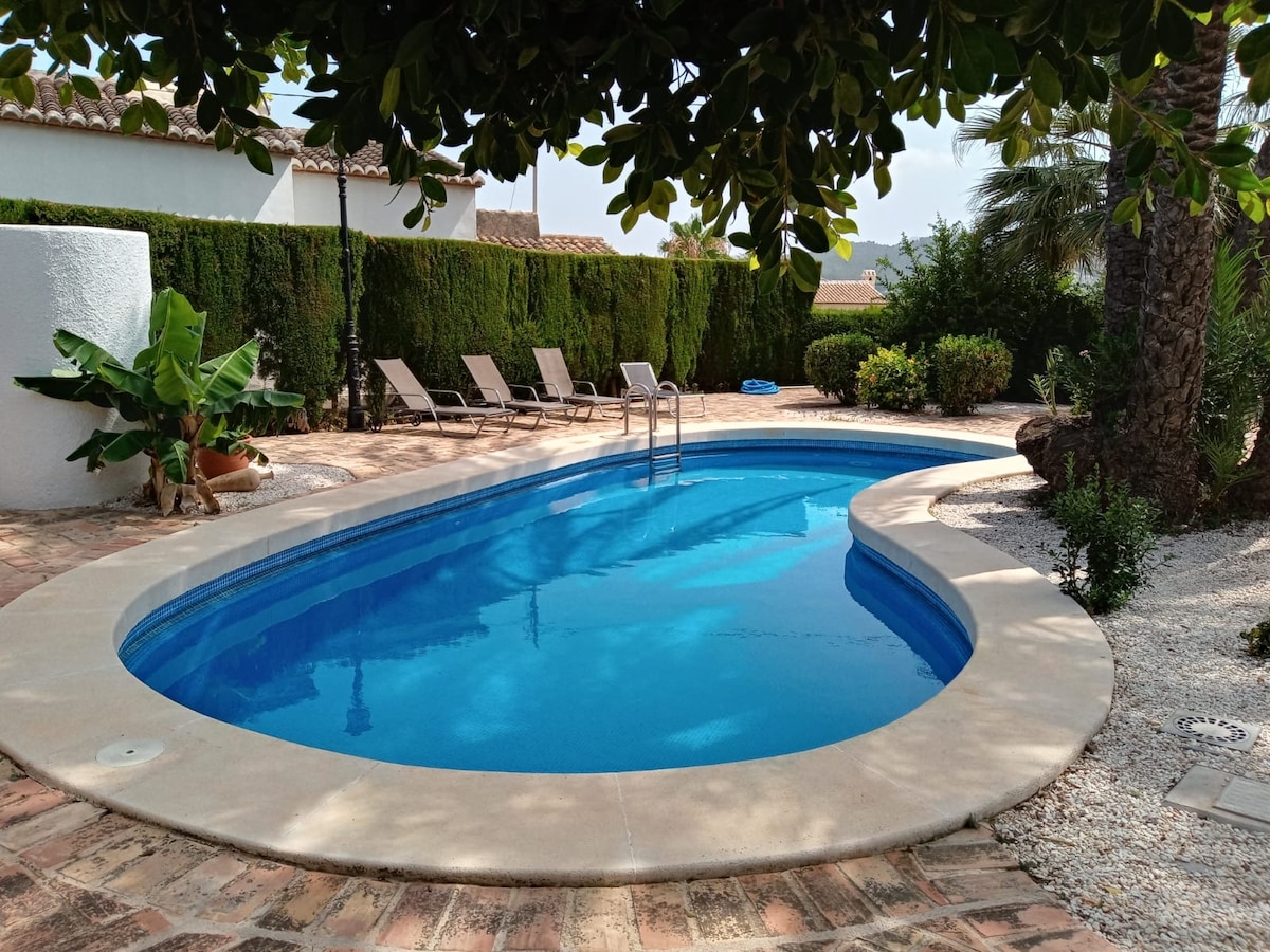 A swimming pool is surrounded by a landscaped garden, featuring palm trees and neat hedges. Loungers are positioned alongside the pool for relaxation. The pool’s clear blue water reflects the natural surroundings, with a sunbathing area paved with terracotta tiles.