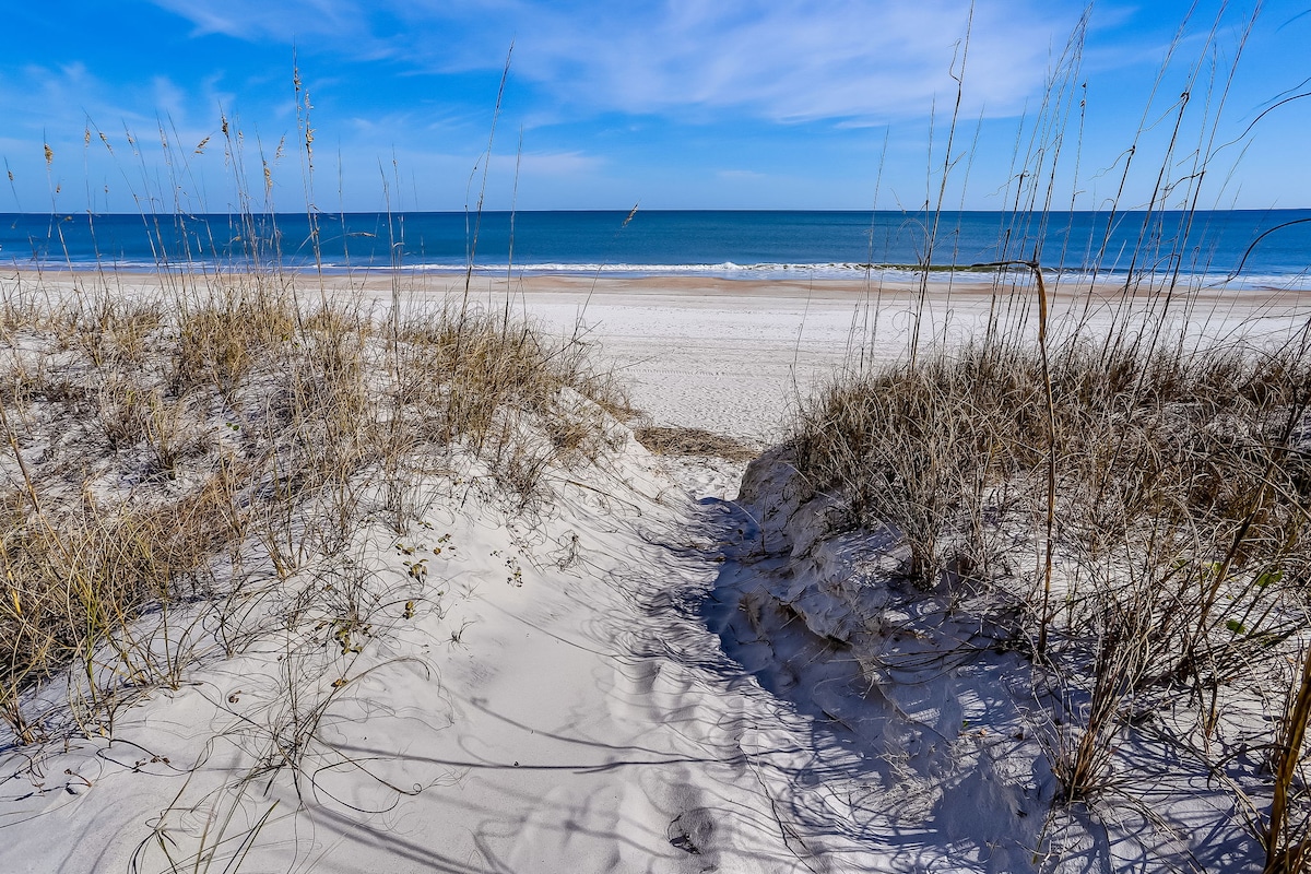 A sandy pathway leads through coastal grasses to the beach, where the gentle waves meet the shore. The horizon showcases the calm blue water under a clear sky, creating a serene seaside atmosphere.