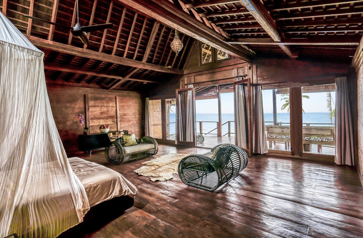 A spacious bedroom features traditional wooden architecture with high ceilings and rich wooden flooring. Large glass doors offer a view of the ocean. A cozy bed is complemented by woven seating arrangements, and the room is softly illuminated by natural light.