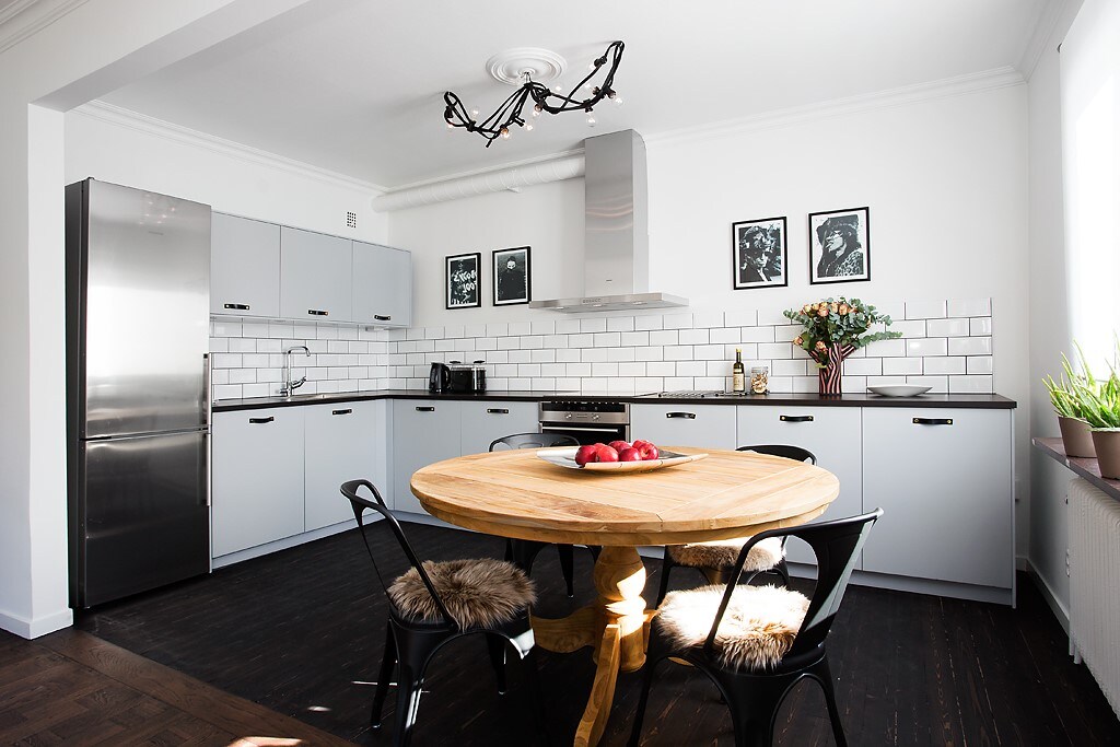 A modern kitchen is presented with light grey cabinetry and a spacious circular wooden table at the center. Black chairs surround the table, which is adorned with a bowl of apples. A stainless steel refrigerator and a stainless steel hood above the stove are visible. Minimalist wall art complements the clean design.