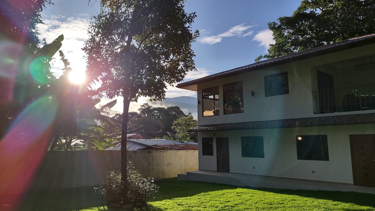 The exterior of a two-story building is shown, featuring large windows and a spacious lawn. Sunlight filters through the trees, casting soft reflections on the grass. The surrounding nature enhances the serene atmosphere, providing a sense of tranquility.