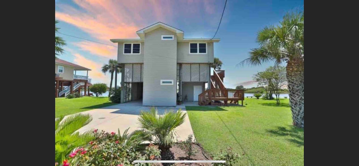A two-story house is situated on a large grassy yard, featuring palm trees and colorful flower beds at the front. The structure has multiple windows and a staircase leading to the entrance. A serene sky is visible, blending pink and blue hues.