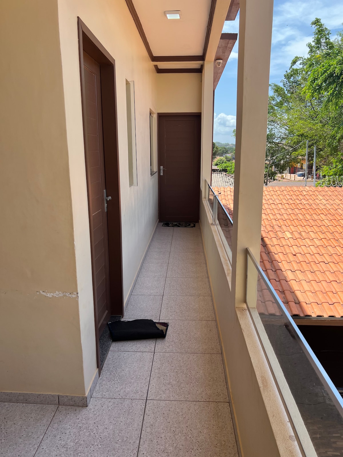 A hallway is depicted, featuring light-colored walls and tiled flooring. Two closed doors are visible along one side, with a narrow space between them. Natural light enters through a nearby window, while a section of the tiled floor includes a thin mat.