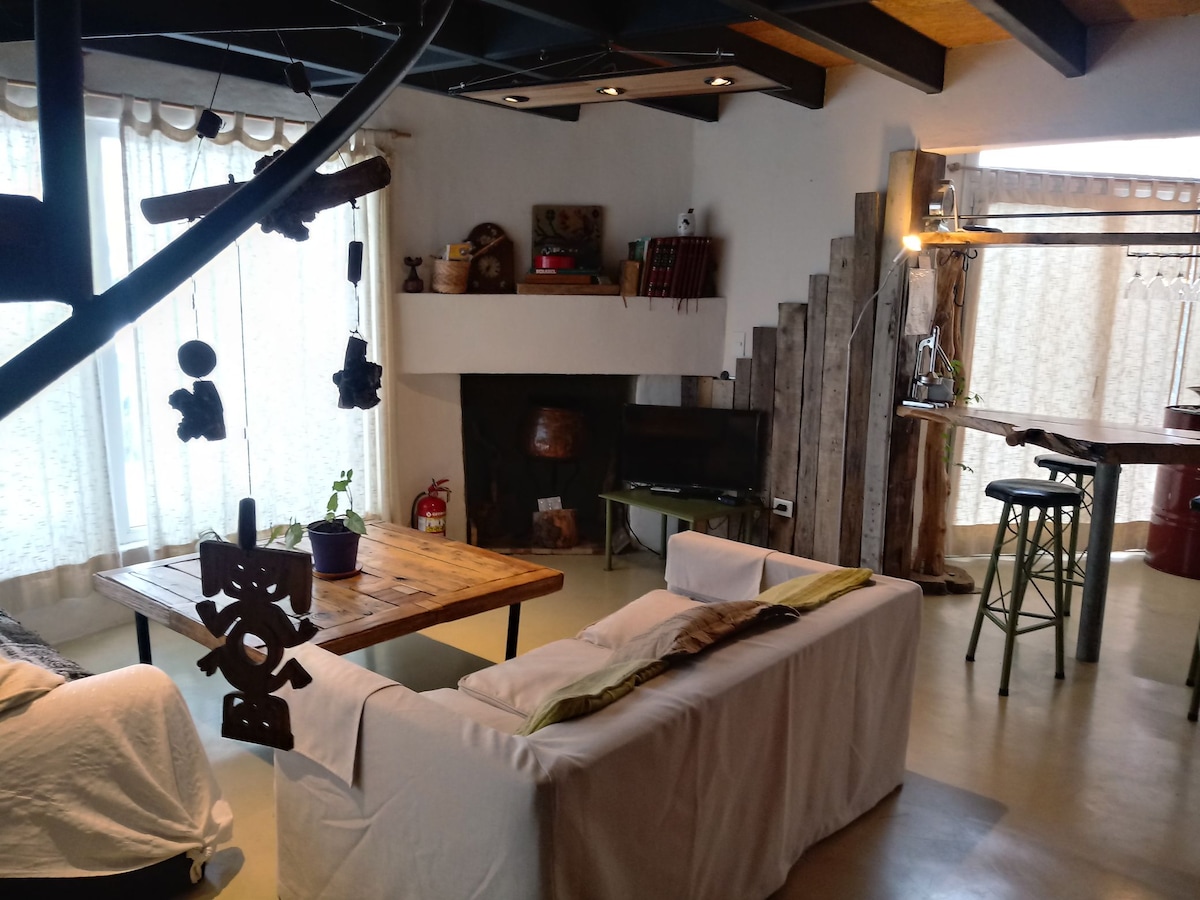 An open living area is featured, with natural light streaming through large windows. A rustic wooden table sits at the center, surrounded by a white sofa. A fireplace and a television are positioned nearby, while a high-top table adds a casual dining option against a backdrop of textured walls.