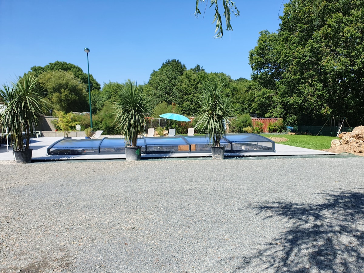 An outdoor area features a covered swimming pool surrounded by greenery. Sun loungers and an umbrella are positioned nearby on a patio. The landscape includes various plants and trees, providing a serene environment for relaxation.