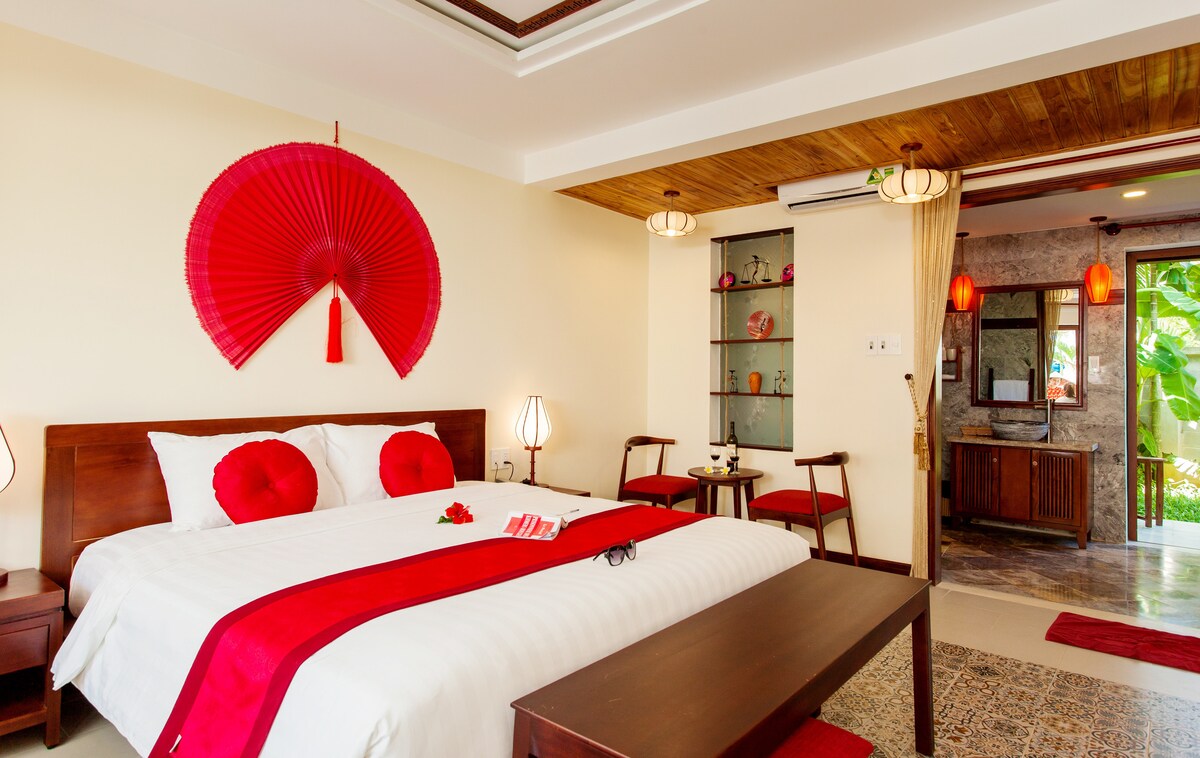 A spacious room is decorated with a large red fan on the wall above the bed. Two round red cushions are placed on the white bedding. Wooden furniture includes a table and chairs, creating a cozy seating area. Soft lighting adds to the inviting atmosphere.