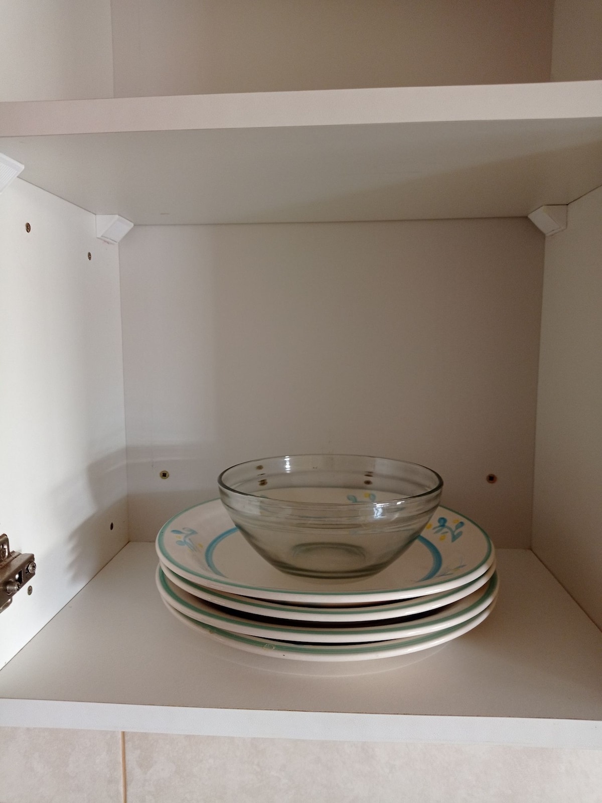 A clean, white kitchen cabinet is shown with neatly arranged dishware. A stack of several dinner plates is positioned at the bottom, topped by a clear glass bowl. The interior is well-lit, showcasing the simplicity and organization of the space.