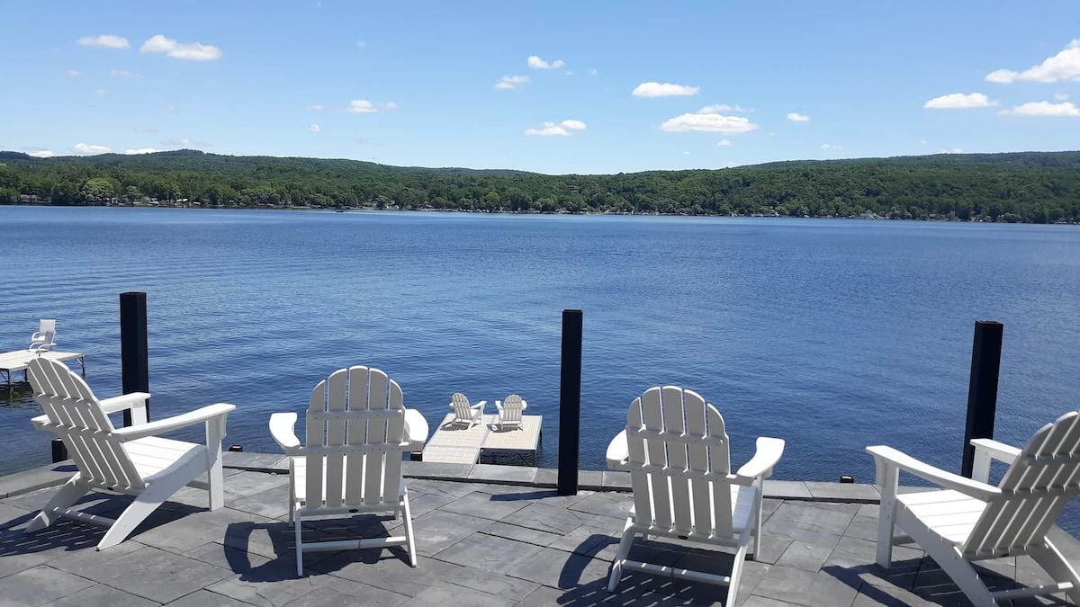 Four white Adirondack chairs are positioned on a stone patio, facing a calm lake with clear blue water. Lush green hills are visible in the background under a bright blue sky with scattered clouds.