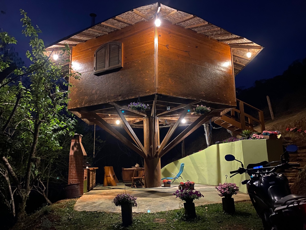 A wooden chalet is elevated on sturdy pillars, illuminated by soft outdoor lighting at night. The surrounding area features decorative planters with flowers, and a barbecue area is visible. A motorcycle stands beside the entrance, and a seating area is positioned nearby.