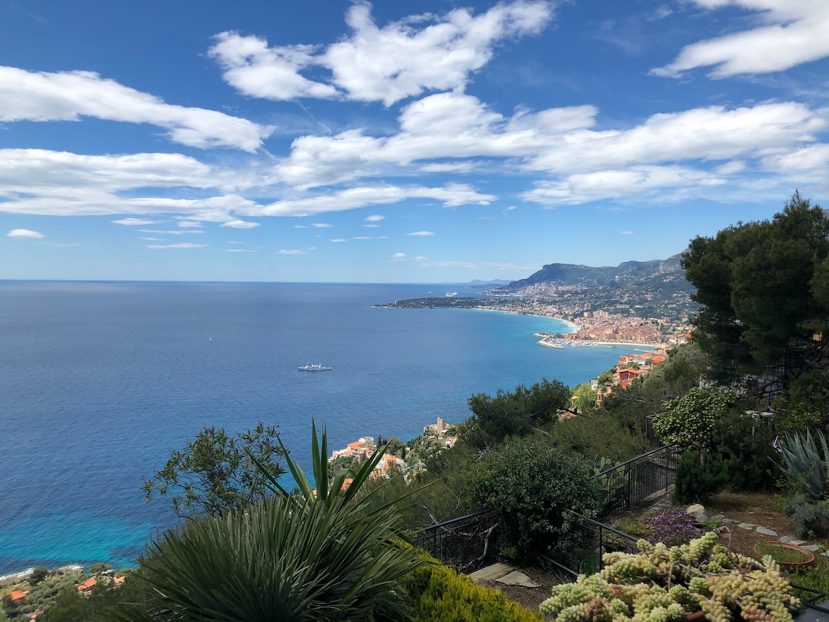 A panoramic view of the coastline is displayed, featuring a vibrant blue sea stretching towards the horizon. The sky contains scattered clouds, while valleys and hills are visible in the distance. Lush greenery, including palm plants and coastal vegetation, frames the scene in the foreground.