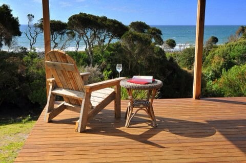 A wooden deck features a comfortable chair facing the ocean, accompanied by a small round table. A glass of wine and an open book rest on the table. Lush greenery surrounds the area, with views of the sea under a clear blue sky.