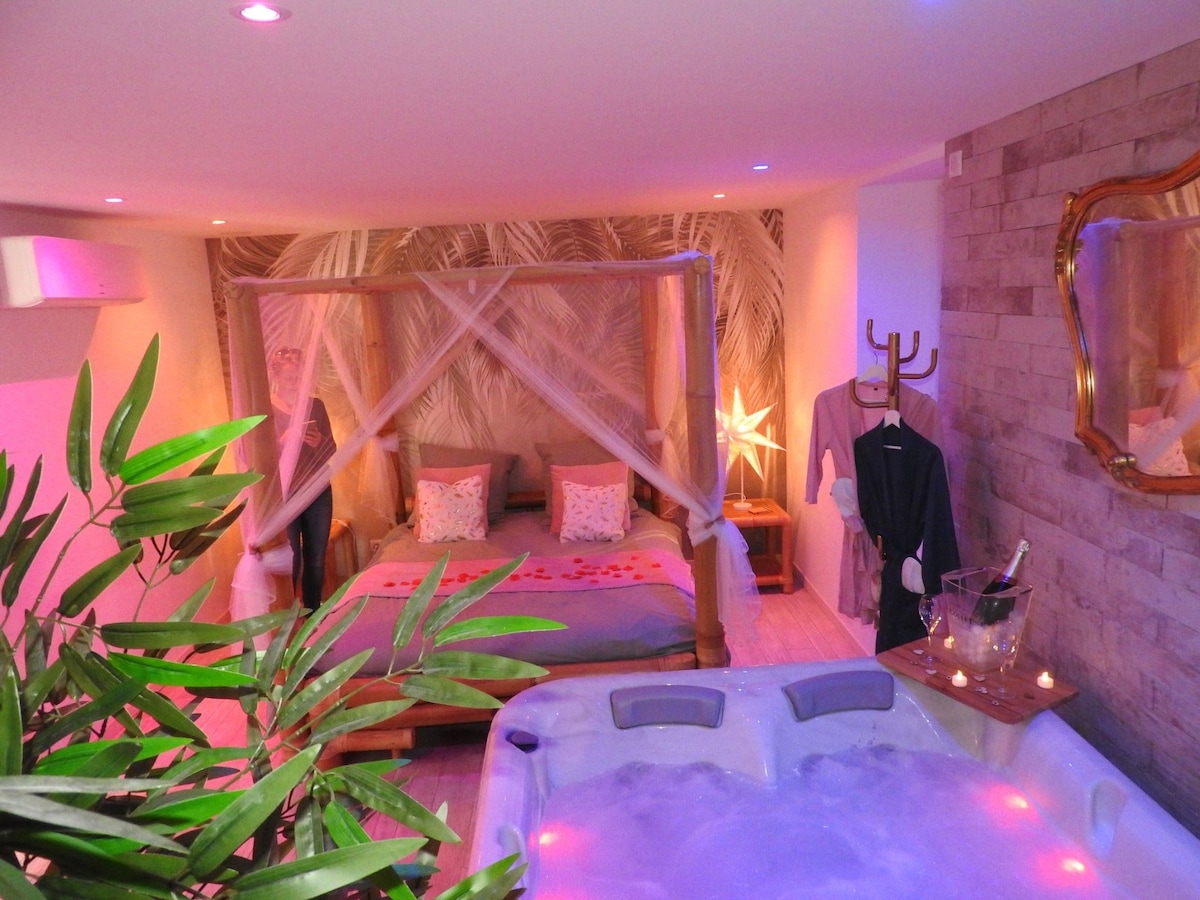 A cozy sleeping area features a spacious bed draped with light, sheer fabric. Soft lighting creates an intimate setting. A private jacuzzi is positioned nearby, accompanied by a small table holding champagne. Tropical-themed decor adds a serene touch to the atmosphere.
