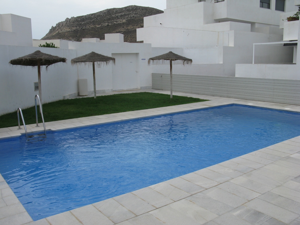 A rectangular pool is set within a paved area, surrounded by green grass. Two thatched umbrellas provide shade near the pool's edge. White buildings create a modern backdrop, with a hillside visible in the distance under an overcast sky.