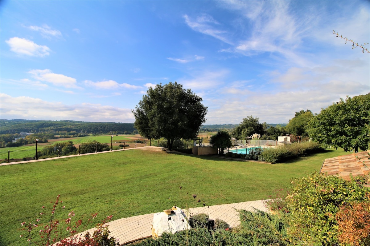 A panoramic view captures a lush green lawn leading to a shared heated pool. The landscape stretches towards distant rolling hills under a partly cloudy sky, enhanced by natural trees dotting the property.