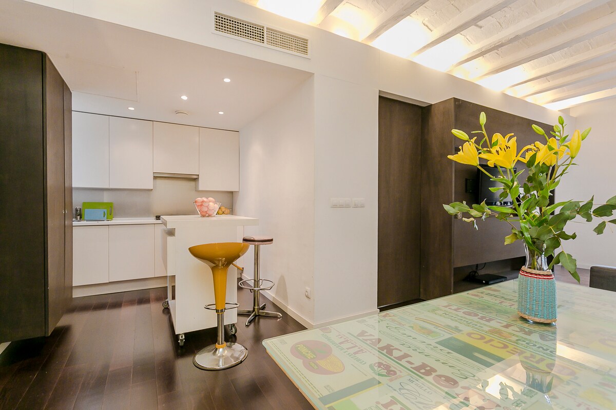 An open kitchen and living area are highlighted, featuring sleek white cabinetry and modern appliances. A small dining table with a decorative vase of flowers sits to the right. Natural light brightens the space, complemented by wooden beams on the ceiling.