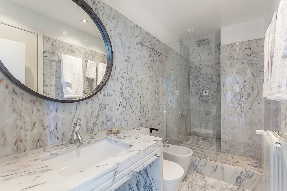 A sleek bathroom is presented, featuring white Portuguese marble walls and surfaces. A round mirror hangs above a modern sink, with a bidet and toilet positioned nearby. A spacious shower enclosure showcases transparent glass, enhancing the room's openness and contemporary design.