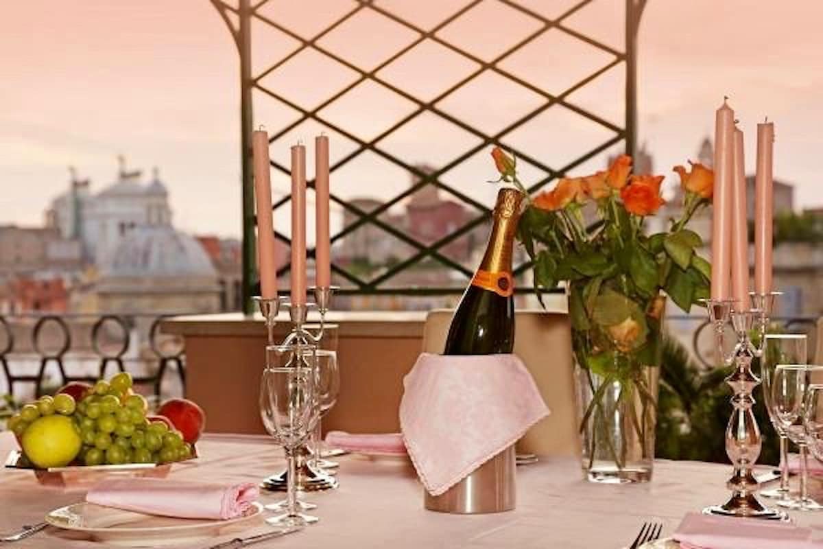 A dining table is set on a terrace, featuring a silver ice bucket with a bottle of champagne. Surrounding are elegant glassware, pink candles, and a vibrant bouquet of roses. Fresh fruit, including grapes and apples, is displayed, with a backdrop of the Roman skyline at sunset.