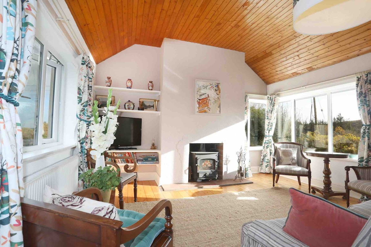 A bright lounge area features high wooden ceilings and large windows providing abundant natural light. A wood-burning stove is positioned below a TV unit, while a round table and comfortable seating create a welcoming space for relaxation. Decorative plants add a touch of nature.