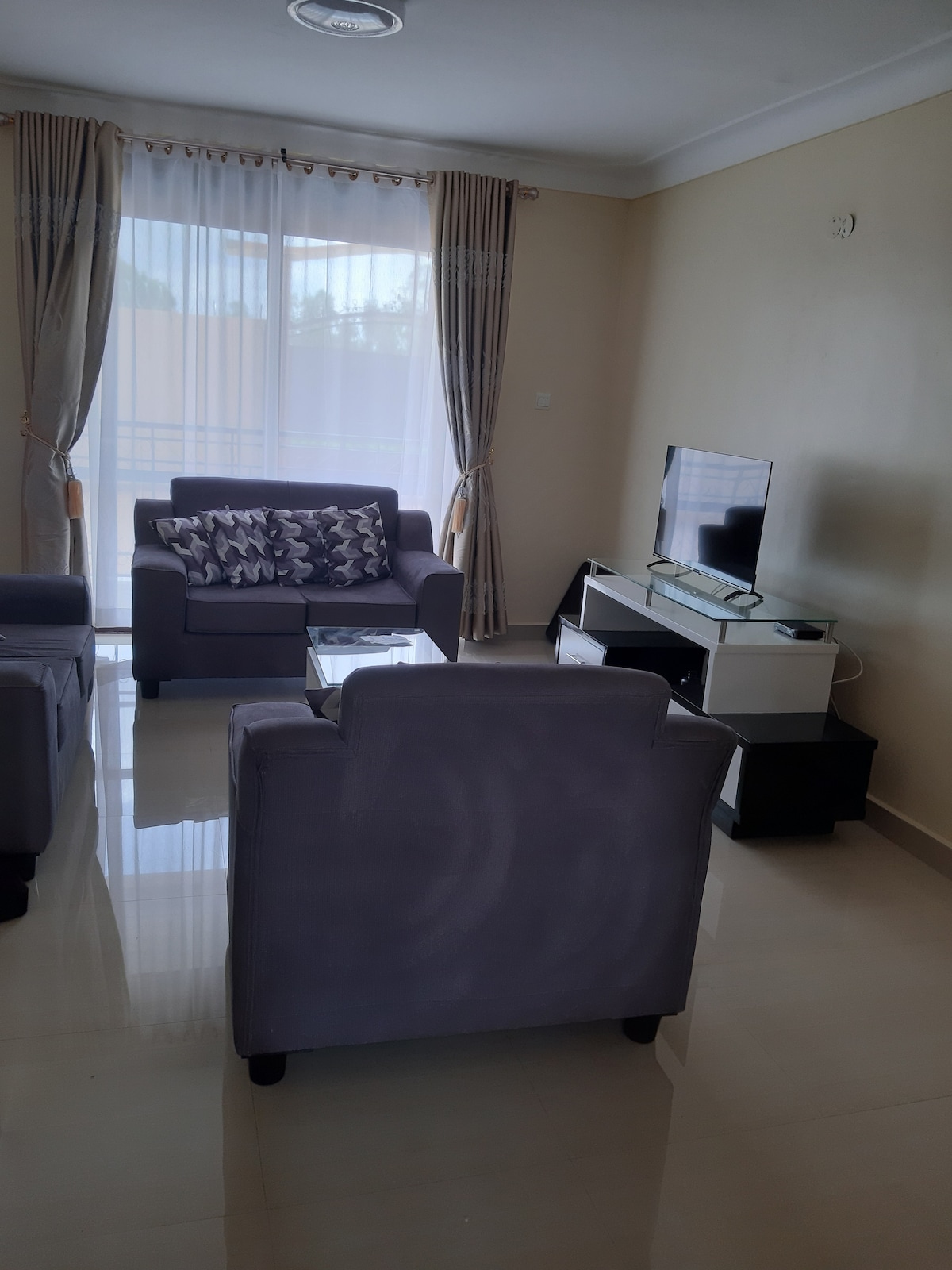 A bright living area features two purple armchairs and a cozy couch adorned with patterned cushions. Large windows with sheer curtains allow natural light to fill the space. A television is positioned on a white and black console, creating a welcoming atmosphere.